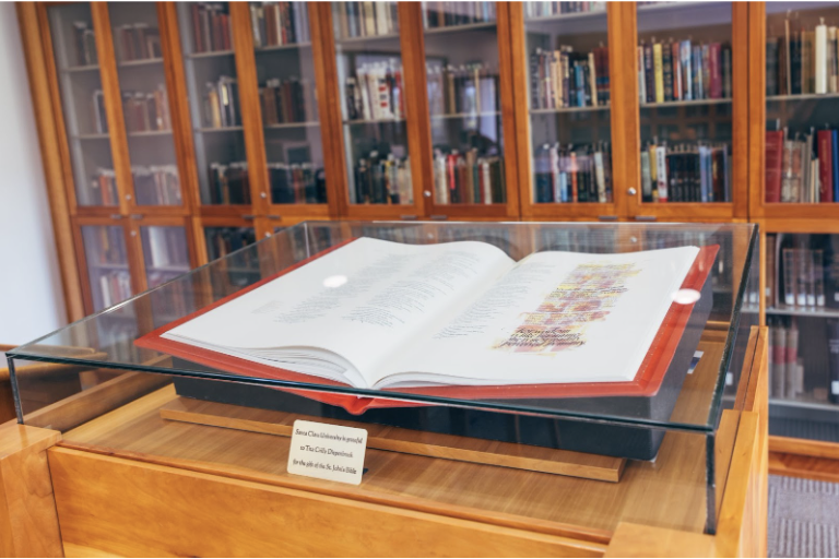 A picture of the special collections room. In the foreground is a large book in a glass case. In the background is a wall of glass cabinets with books inside. Credit: Photo by Miguel Ozuna.